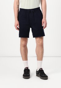 Marineblå shorts i et mykt, lett stoff med elastisk midje og snøring. Brukt med hvite sokker og svarte joggesko.
