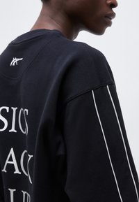 Κοντινή λήψη ενός ατόμου που φοράει μαύρο φούτερ με λευκό κείμενο και λεπτές λευκές ρίγες κατά μήκος του μανικιού.