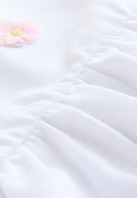 Hvidt stof med et plisseret design og en lille lyserød blomst som accent. Teksturen virker blød og glat og fremhæver de delikate detaljer.