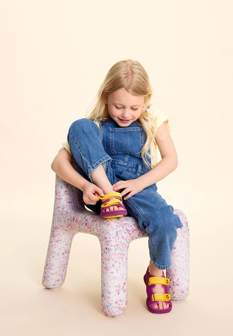 Sandali viola con cinturini gialli ai piedi di un bambino, seduto su una sedia colorata, morbida e testurizzata, indossando una salopette di jeans e una maglietta leggera.
