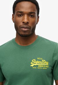 Camiseta de algodón verde con texto bordado amarillo que dice "REAL SuperDry," junto con caracteres japoneses y un lema.