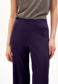 Fialové žebrované kalhoty s vysokým pasem. Textura látky je měkká s jemným vertikálním vzorem. Spárováno s pruhovaným topem, vystaveno na neutrálním pozadí.