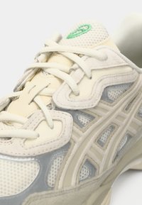 Chaussure de sport ivoire et grise avec des textures en maille et en daim, présentant un logo vert, un design de superposition complexe et un système de laçage.