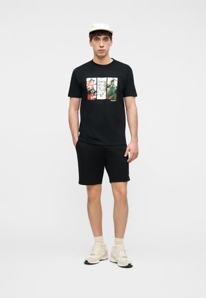 Mladi moški stoji ob preprostem ozadju, oblečen v črno majico z grafičnim potiskom, črne kratke hlače, bele superge, nogavice in belo kapo.