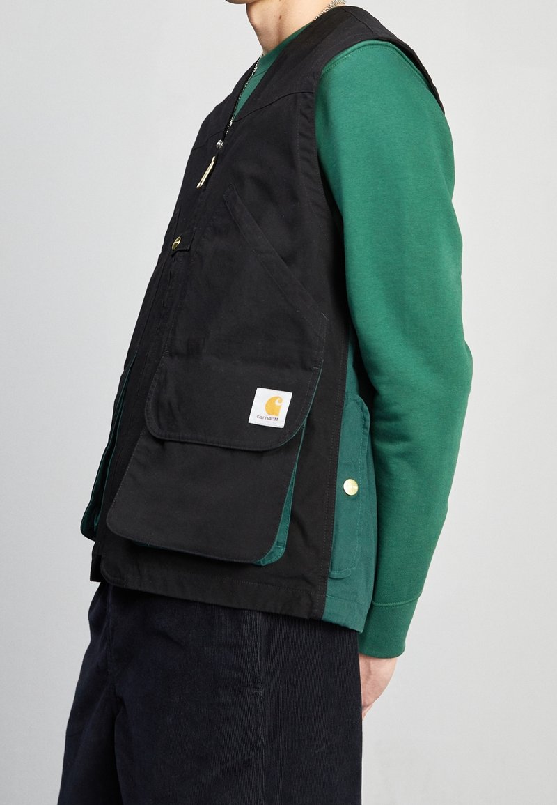 Schwarze Weste mit großen Fronttaschen, goldenen Beschlägen und einem Carhartt-Logo. Über einem grünen Sweatshirt getragen, das ein schlichtes Design und robusten Stoff hat.