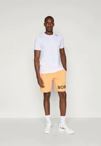 Λευκή μπλούζα και πορτοκαλί σορτς με την ένδειξη "BOR" τυπωμένη στα μαύρα. Το σύνολο διαθέτει χαλαρή γραμμή, απαλή ύφανση και αντίθετα χρώματα.