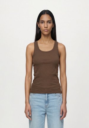 Jeune femme aux longs cheveux foncés portant un haut marron sans manches et un jean bleu clair, debout devant un fond clair uni.