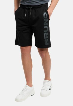 Mann trägt schwarze Shorts mit Kordelzug, auf dem linken Bein vertikal bedruckt mit "BRUNO", und weiße Sneakers mit schwarzem Text an den Außenseiten.