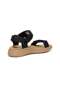 Mustat kimaltelevat sandaalit säätövaroilla, litteä beige pohja ja pehmustettu pohjallinen. Sivukuva, joka korostaa hihnasuunnittelua ja tekstuuria.