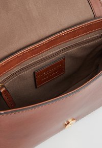Interno di una borsa in pelle marrone con una fodera in tela beige, una tasca con zip e un’etichetta in pelle con un marchio impresso.