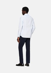 Camisa de color azul claro con yugo trasero y dobladillo redondeado, combinada con pantalones oscuros de color navy. La camisa tiene mangas largas y cuello abotonado, llevada con zapatos negros.