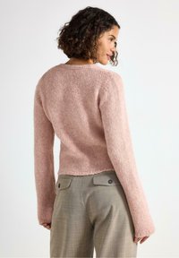 Kvinna med lockigt hår iförd en mjuk ljusrosa stickad tröja och beige skräddarsydda byxor, sedd bakifrån mot en enkel bakgrund.
