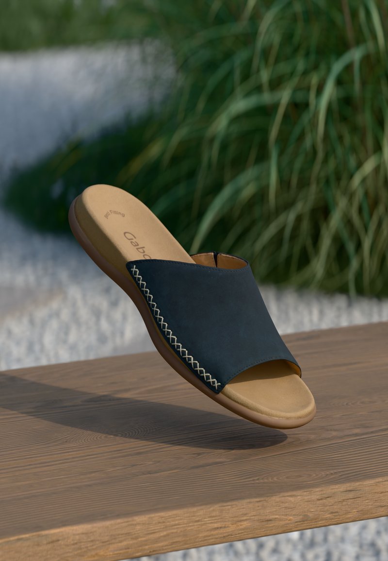 Braune Sohlen-Slide-Sandale mit schwarzem Obermaterial und weißen Seitennähten, auf einer Holzoberfläche präsentiert, mit unscharfen grünen Pflanzen im Hintergrund.