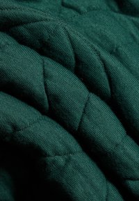 Steppelt zöld anyag, puha textúrával, varrott mintákkal és réteges hajtásokkal, amelyek tapintásra kellemes és dús megjelenést kölcsönöznek.