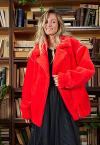 Piros műszőrme kabát széles gallérral, cipzáros záródással és túlméretezett ujjakkal, fekete tüll szoknyával párosítva, könyvespolc háttér előtt.