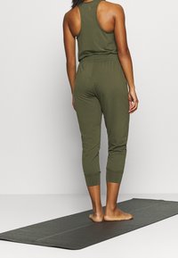 Combinaison vert olive à manches sans manches, jambes fuselées longueur cheville, texture douce, portée pieds nus sur un tapis de yoga noir.