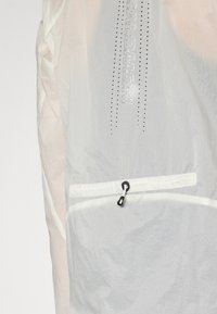 Lightweight, semi-transparent jacket in white, featuring a grid texture, small front pocket with black toggle, and "SALOMON" logo in silver.