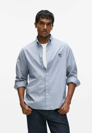 Jeune homme portant une chemise à carreaux bleu clair à boutons avec manches retroussées et un jean foncé, debout les mains partiellement dans les poches.