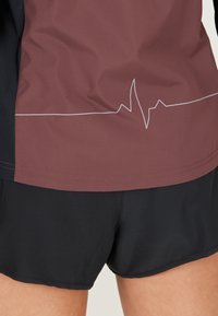 Bordowy i czarny sportowy top z białym motywem linii tętna; charakteryzuje się gładką teksturą i kontrastującymi kolorami. Czarne shorty poniżej.