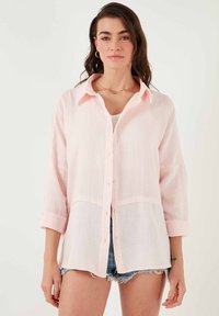 Light pink button-up shirt with a textured fabric, long sleeves rolled at the cuffs, and a white bottom panel. Shorter warm-weather style.
