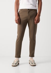 Pantalones marrones ajustados con una textura suave, que cuentan con bolsillos laterales y un cierre de botón único, combinados con zapatillas verdes y beiges.
