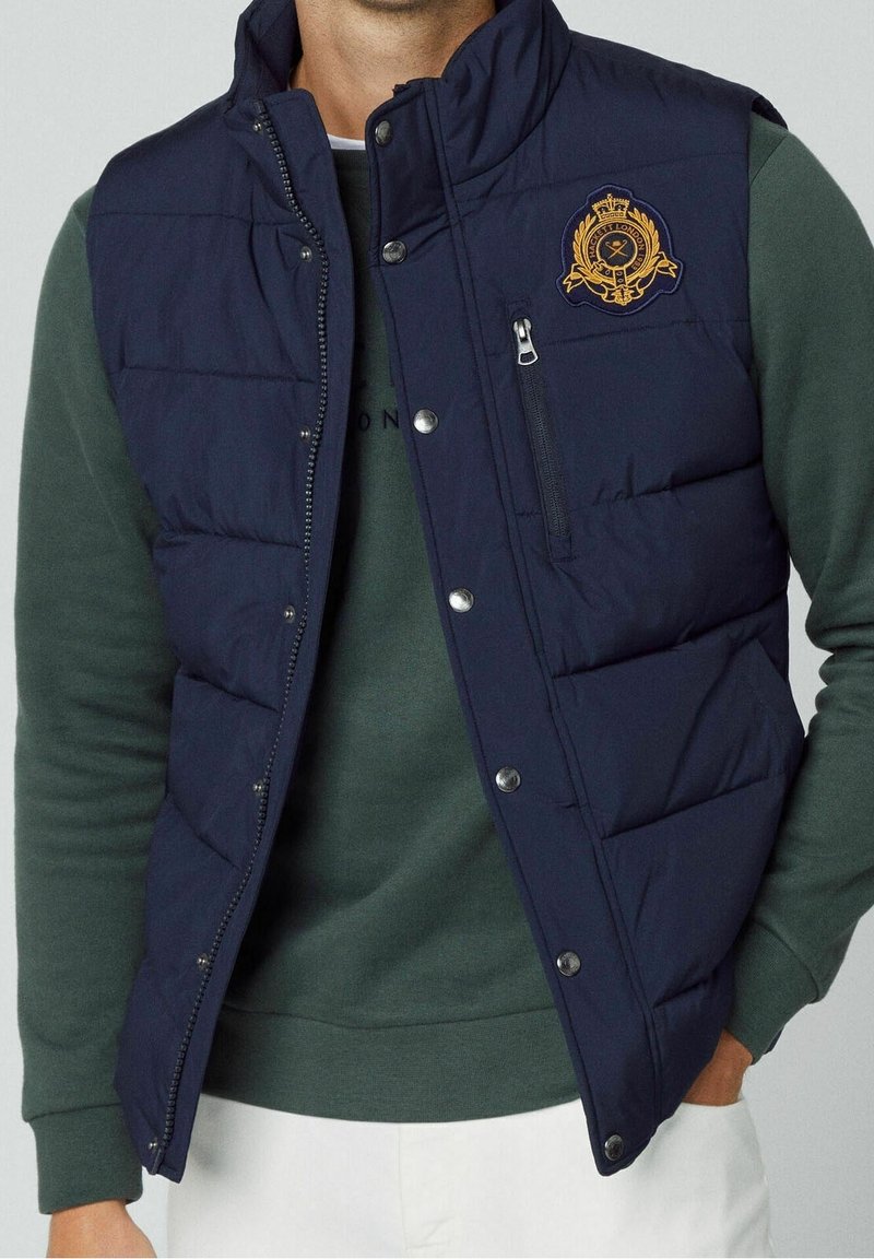 Homme portant un gilet matelassé bleu marine avec fermeture éclair et boutons-pression sur un sweat-shirt vert et un pantalon blanc, avec un blason brodé sur la poitrine.