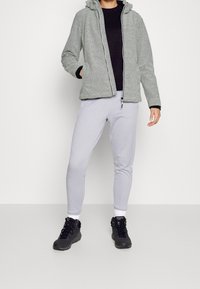 Graue strukturierte Zip-Hoodie-Jacke über einem schwarzen Top, kombiniert mit hellgrauen schmal geschnittenen Jogginghosen und schwarzen Wanderschuhen.