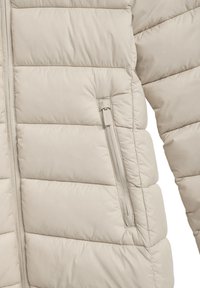 Chaqueta acolchada beige con textura de acolchado horizontal, bolsillo lateral con cremallera y tejido suave. Cierre frontal con cremallera y diseño minimalista.