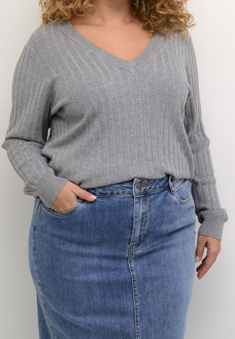 Personne portant un pull gris côtelé à col en V rentré dans une jupe en denim bleue taille haute, avec une main dans la poche de la jupe.