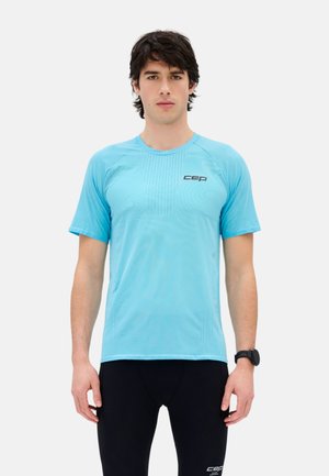 Junger Mann trägt ein hellblaues kurzärmeliges Sportshirt und schwarze Hose, mit einer schwarzen Smartwatch am linken Handgelenk, steht frontal.