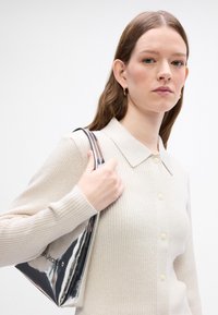 Beige ribbet cardigan med kragehals og knappelukking. Modellen holder en blank sølvveske med glidelås.