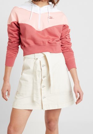 Vrouw draagt een roze-witte cropped Nike hoodie gecombineerd met een hooggetailleerde crèmekleurige rok met knopen aan de voorkant en een strikceintuur.