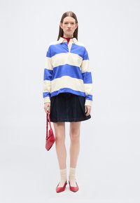 Femme debout portant une chemise à manches longues rayée bleu et blanc, une jupe noire, des chaussettes beiges, des chaussures rouges, et tenant un petit sac à main rouge.