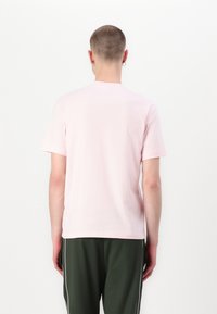 Kurzarm-T-Shirt aus leichtem Baumwollstoff in Hellrosa, mit rundem Ausschnitt, minimalistischem Design, getragen zu dunkelgrünen Hosen mit weißen Seitenstreifen.
