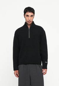 Zwarte zip-neck trui in een gestructureerde brei, met een hoge kraag, geribde manchetten en zoom. Gecombineerd met grijze geplooide broeken.
