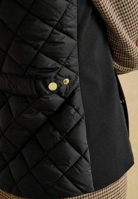 Zbliżenie na czarną pikowaną kurtkę z złotymi zatrzaskami noszoną na ubraniu w wzór houndstooth.