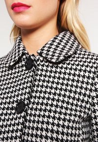 Kabát s černobílým kostkovaným vzorem houndstooth, s límečkem a černými knoflíky, s tkanou texturou a strukturovaným designem.