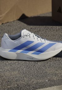 Scarpa da corsa bianca con strisce blu e suola spessa imbottita con etichetta Lightstrike Pro su una superficie esterna ruvida.