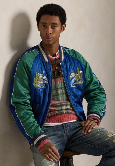 Modro-zelena bomber jakna z vezenim prizorom gora in dreves, s črtastimi manšetami, nošena čez rdečozelen karo pulover. Obrabljene kavbojke.