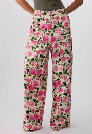 Hose mit weitem Bein und rosa-grünem Blumenmuster, große Front-Cargotaschen, getragen mit olivgrünem Tanktop und weißen Sandalen mit offenem Zehenbereich.