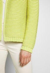 Jasnozielony sweter na drutach z fakturowanym wzorem, ściągaczowymi mankietami i otwartym przodem, w połączeniu z kremowymi spodniami.