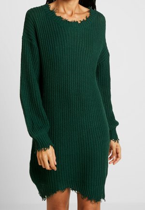 Vrouw draagt een donkergroene, met lange mouwen gebreide trui-jurk met rafelige randen bij de manchetten, halslijn en zoom.