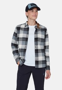 Camicia di flanella a quadri grigia, bianca e nera; caratterizzata da chiusura con bottoni, taschino sul petto, abbinata a una t-shirt bianca e pantaloni blu navy.