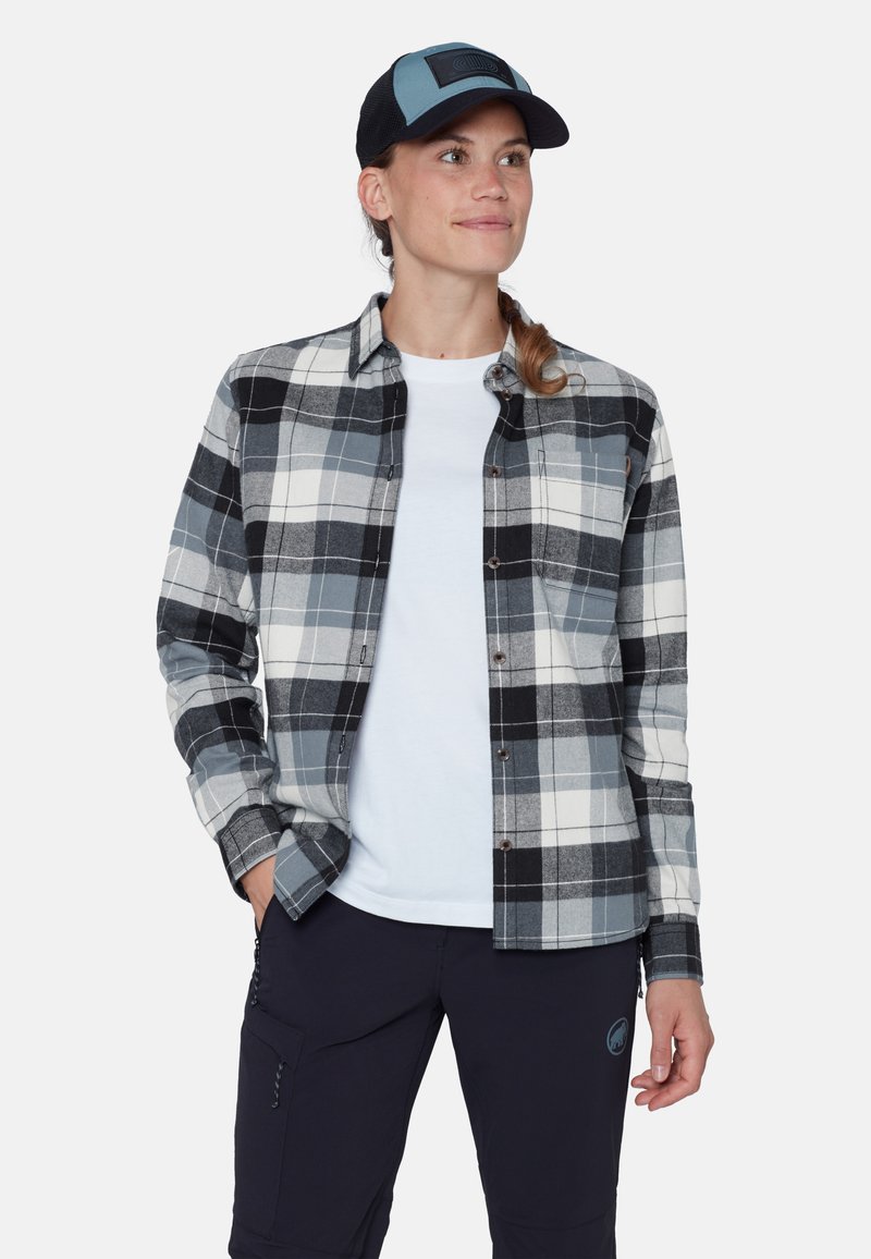 Camicia di flanella a quadri grigia, bianca e nera; caratterizzata da chiusura con bottoni, taschino sul petto, abbinata a una t-shirt bianca e pantaloni blu navy.