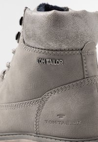 Bottine en cuir gris à surface texturée, coutures robustes et col matelassé. Présente un logo embossé "TOM TAILOR" sur le côté.