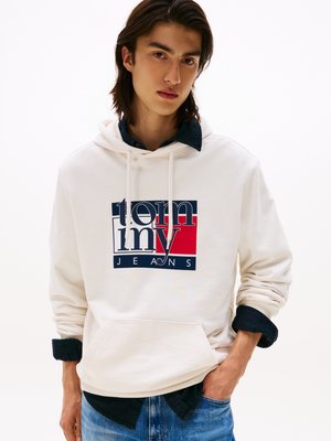 Felpa bianca con cappuccio con una grafica frontale in blu navy e rosso con il testo "tommy jeans". Indossata sopra una camicia in denim scuro, mostrata con una vestibilità rilassata.