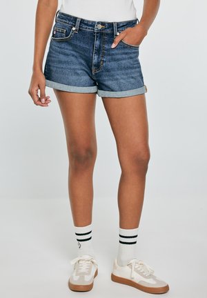 Osoba nosząca dżinsowe szorty z podwiniętym brzegiem, białe trampki z gumową podeszwą, białe skarpetki z czarnymi paskami, stojąca na jednolitym tle.