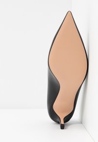 Chaussure noire à talon haut à bout pointu montrée par la semelle, positionnée verticalement contre un mur d’angle blanc.