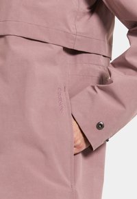 Ljusrosa vattentät jacka med hög krage, tryckknappsstängning och subtilt broderad logotyp på sidan. Slät yta, figursydd passform.