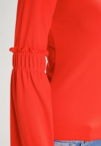 Röd räfflad långärmad topp med samlade ärmslut, med en strukturerad yta och en figurnära siluett. Inga ytterligare dekorationer synliga.